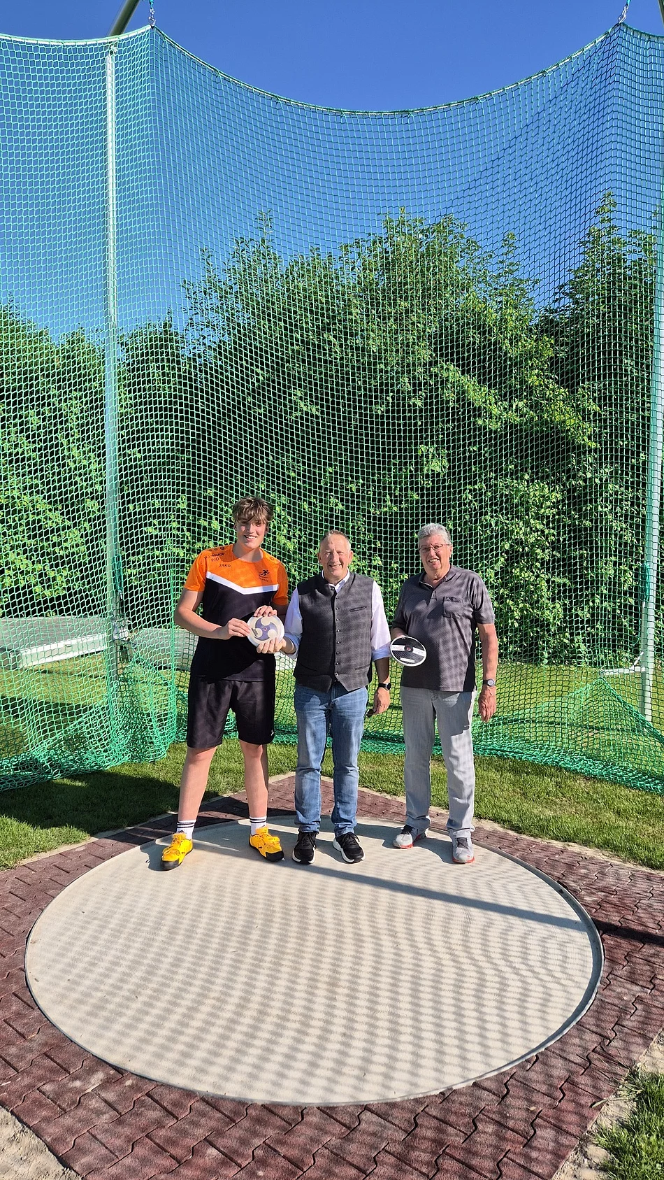 Drei Männer stehen auf einer Diskuswurfanlage vor einem grünen Netz, der linke trägt Sportkleidung und hält einen Diskus in der Hand