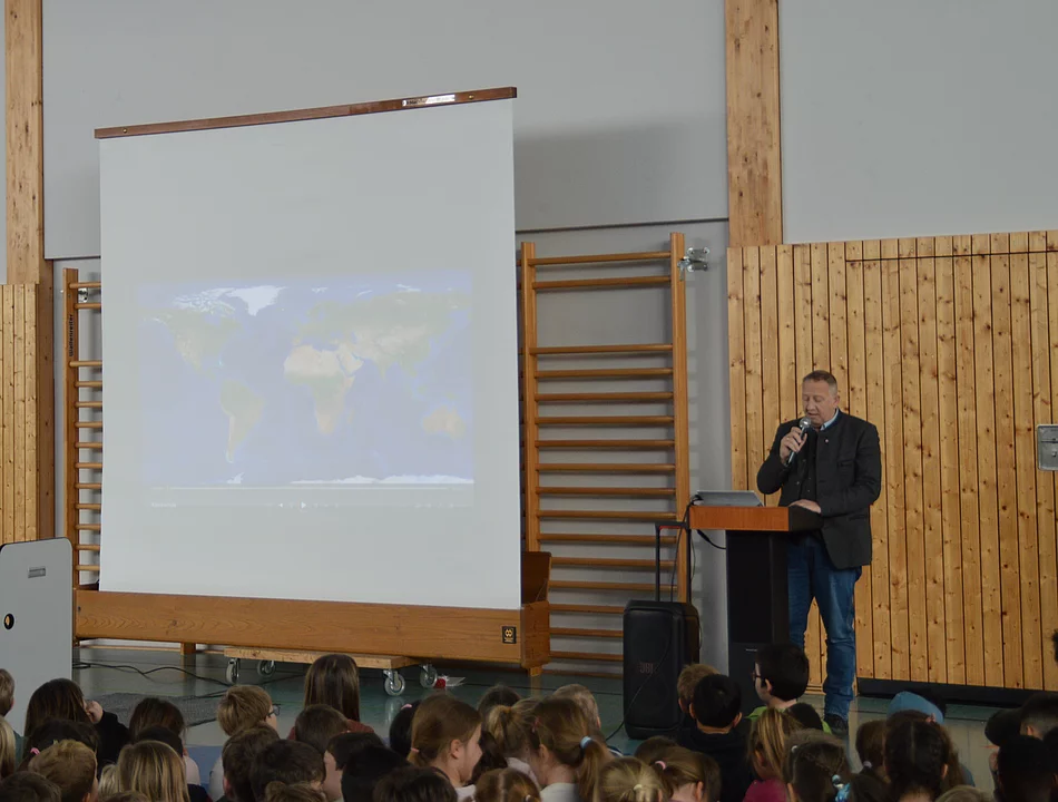 Mann mit Mikrofon steht an Rednerpult vor Leinwand mit Weltkarte, davor sitzende Kinder in Turnhalle mit Holzverkleidung