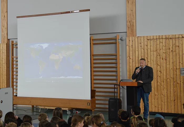 Mann mit Mikrofon steht an Rednerpult vor Leinwand mit Weltkarte, davor sitzende Kinder in Turnhalle mit Holzverkleidung