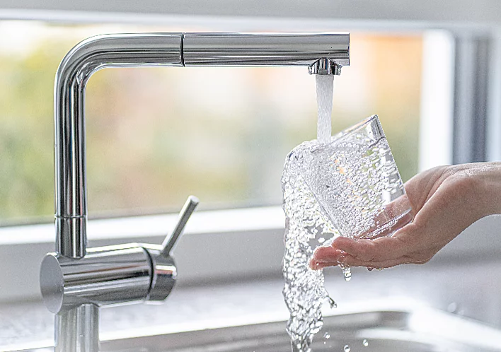 Silberner Wasserhahn mit laufendem Trinkwasser auf weibliche Hand, die ein Wasserglas hält und es befüllt
